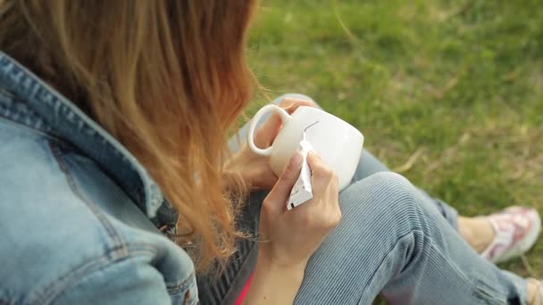 Close-up of female hands drawing flowery pattern on a white cup. Girl paints ornament sitting on the grass in the evening. — Stock Video