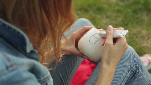 Gros plan des mains féminines dessinant un motif fleuri sur une tasse blanche. Fille peint ornement assis sur l'herbe dans la soirée . — Video