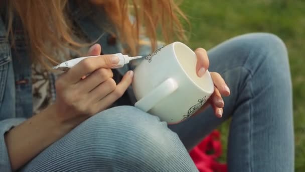 Gros plan des mains féminines dessinant un motif fleuri sur une tasse blanche. Fille peint ornement assis sur l'herbe dans la soirée . — Video