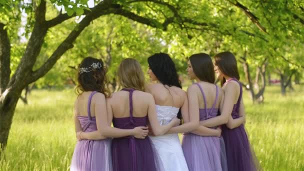 Mira por detrás de la novia emocional y damas de honor que hablando y sonriendo. Caucásico niñas en morado vestidos posando al aire libre . — Vídeo de stock