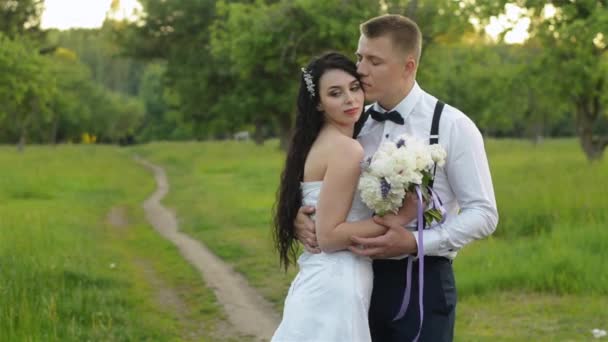 Fashionable young wedding couple enjoy each other in a park or garden standing on the road in the evening. Stylish newlyweds in hipster clothes hugging and looking into the distance. — Stock Video