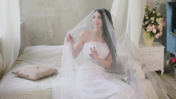 The morning of a gentle attractive brunette. Beautiful bride is smiling and looking out the window touching the veil while sitting on the bed. — Stock Video