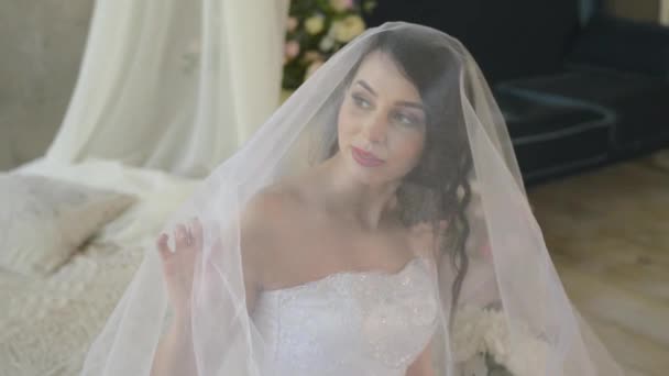 The morning of a gentle attractive brunette. Beautiful bride is smiling and looking out the window touching the veil while sitting on the bed. — Stock Video