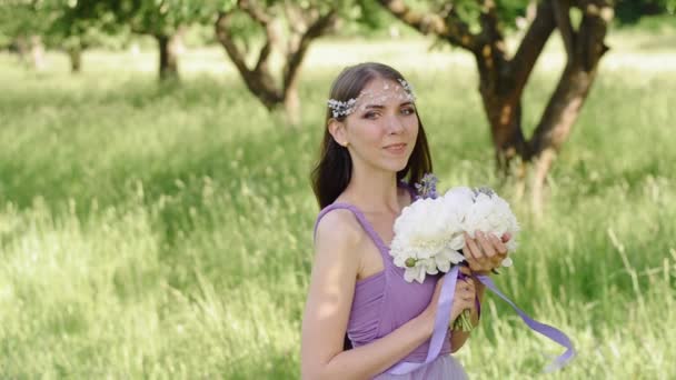 Porträtt av en vacker flicka med professionell make-up. Kaukasiska brunett kvinna poserar och tittar på kameran i lila eller lila klänning i grön trädgård nära frukt äppelträd. — Stockvideo