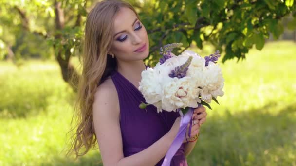 Портрет красивой девушки с ярким макияжем. Кавказская блондинка с букетом цветов позирует и смотрит в камеру в сиреневом или фиолетовом платье в саду . — стоковое видео
