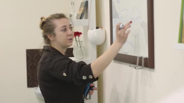 Professional make-up artist teaches the techniques of make-up on a masterclass. Young woman in black shirt draws eyerbrows or something else on glass board with marker. — Stock Video