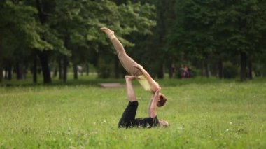 Güzel çift Acro yoga uygulamak. Yeşil çimenlerin üzerinde bir şehir parkta genç yoga eğitmenleri pratik. İki başarılı gençler acro yoga egzersizleri yapın. Erkek ve kadın gün batımında yoga öğrenmek.