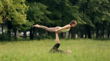 Güzel çift Acro yoga uygulamak. Yeşil çimenlerin üzerinde bir şehir parkta genç yoga eğitmenleri pratik. İki başarılı gençler acro yoga egzersizleri yapın. Erkek ve kadın gün batımında yoga öğrenmek.