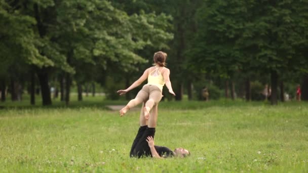 Hermosa pareja practicando acro yoga. Jóvenes instructores de yoga practican en un parque de la ciudad sobre hierba verde. Dos jóvenes exitosos realizan ejercicios de acro yoga. Hombre y una mujer aprenden yoga al atardecer . — Vídeo de stock