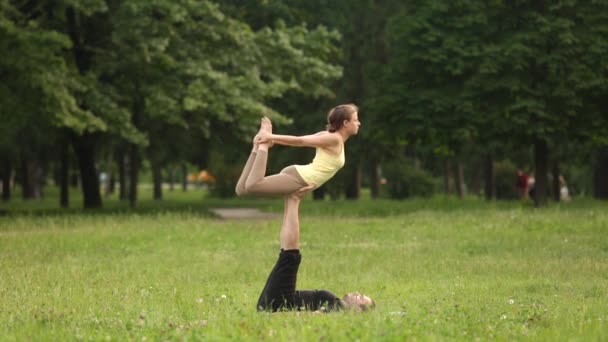 Belo casal praticando acro ioga. Jovens instrutores de ioga praticam em um parque da cidade na grama verde. Dois jovens bem sucedidos realizam exercícios de acro ioga. Homem e uma mulher aprendem ioga ao pôr do sol . — Vídeo de Stock