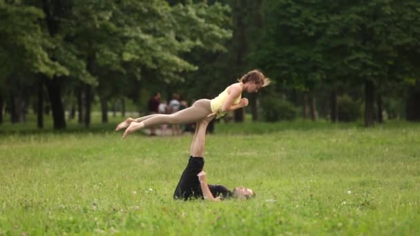 Beau couple pratiquant l'acro yoga. Jeunes instructeurs de yoga pratiquent dans un parc de la ville sur l'herbe verte. Deux jeunes qui réussissent effectuent des exercices d'acro yoga. Homme et une femme apprennent le yoga au coucher du soleil . — Video
