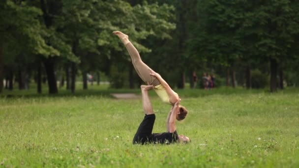 Hermosa pareja practicando acro yoga. Jóvenes instructores de yoga practican en un parque de la ciudad sobre hierba verde. Dos jóvenes exitosos realizan ejercicios de acro yoga. Hombre y una mujer aprenden yoga al atardecer . — Vídeos de Stock