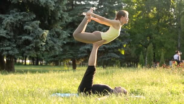 Hermosa pareja practicando acro yoga. Jóvenes instructores de yoga practican en un parque de la ciudad sobre hierba verde. Dos jóvenes exitosos realizan ejercicios de acro yoga. Hombre y una mujer aprenden yoga al atardecer . — Vídeos de Stock