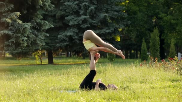 Belo casal praticando acro ioga. Jovens instrutores de ioga praticam em um parque da cidade na grama verde. Dois jovens bem sucedidos realizam exercícios de acro ioga. Homem e uma mulher aprendem ioga ao pôr do sol . — Vídeo de Stock