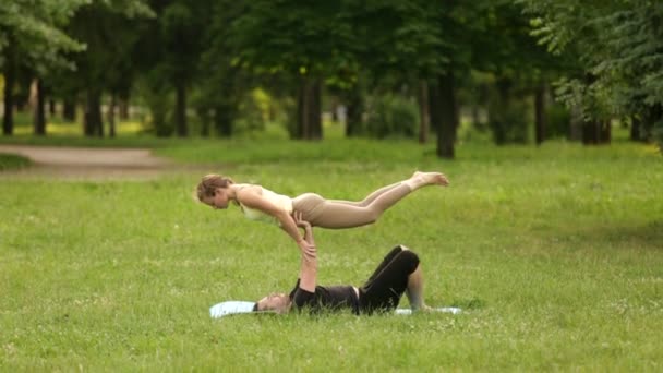 Hermosa pareja practicando acro yoga. Jóvenes instructores de yoga practican en un parque de la ciudad sobre hierba verde. Dos jóvenes exitosos realizan ejercicios de acro yoga. Hombre y una mujer aprenden yoga al atardecer . — Vídeos de Stock