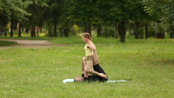 Hermosa pareja practicando acro yoga. Jóvenes instructores de yoga practican en un parque de la ciudad sobre hierba verde. Dos jóvenes exitosos realizan ejercicios de acro yoga. Hombre y una mujer aprenden yoga al atardecer . — Vídeos de Stock