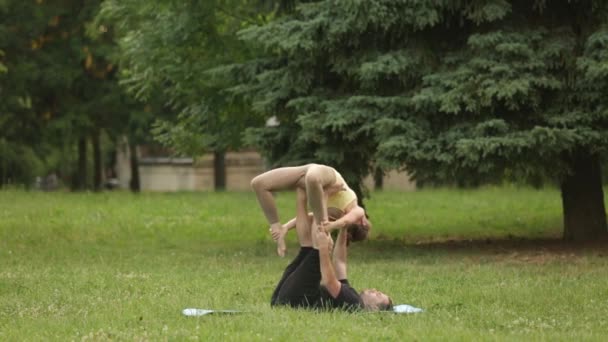 Krásný pár cvičení acro jógy. Mladí instruktoři jógy v městském parku na zelené trávě. Dva mladí a úspěšní lidé provádět cvičení jógy acro. Muž a žena naučit Jóga při západu slunce. — Stock video