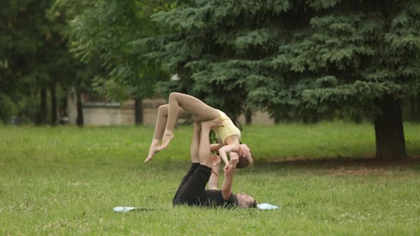 Hermosa pareja practicando acro yoga. Jóvenes instructores de yoga practican en un parque de la ciudad sobre hierba verde. Dos jóvenes exitosos realizan ejercicios de acro yoga. Hombre y una mujer aprenden yoga al atardecer . — Vídeos de Stock