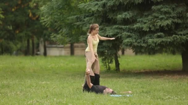 Bella coppia praticare acro yoga. Giovani istruttori di yoga praticano in un parco cittadino su erba verde. Due giovani di successo eseguono esercizi di acro yoga. Uomo e donna imparano lo yoga al tramonto . — Video Stock