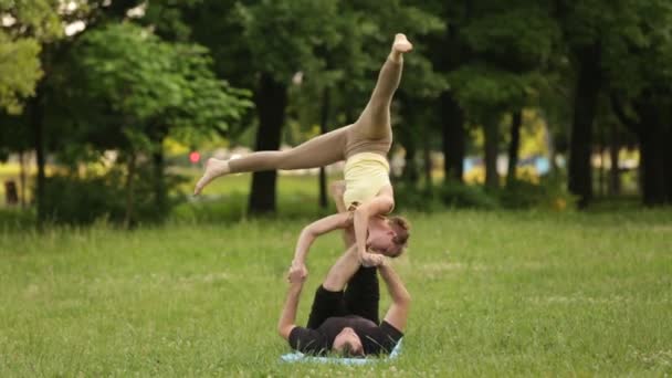 Vackra par öva acro yoga. Unga yoga instruktörer praxis i en park på grönt gräs. Två framgångsrika unga människor utföra acro yogaövningar. Man och en kvinna lär dig yoga i solnedgången. — Stockvideo