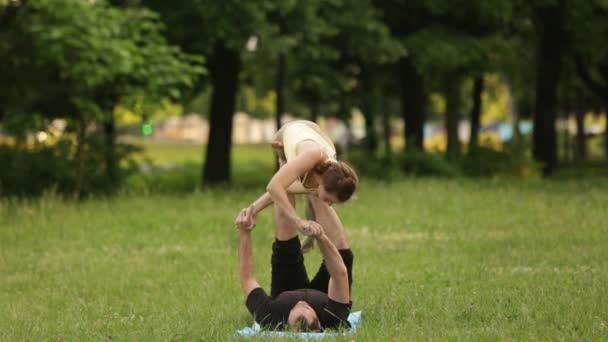 Hermosa pareja practicando acro yoga. Jóvenes instructores de yoga practican en un parque de la ciudad sobre hierba verde. Dos jóvenes exitosos realizan ejercicios de acro yoga. Hombre y una mujer aprenden yoga al atardecer . — Vídeos de Stock