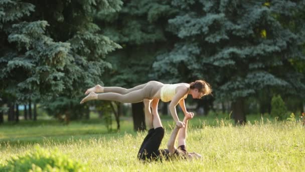 Прекрасная пара, практикующая акро-йогу. Молодые инструкторы йоги практикуют в городском парке на зеленой траве. Два успешных молодых человека выполняют упражнения по акро-йоге. Мужчина и женщина учатся йоге на закате . — стоковое видео