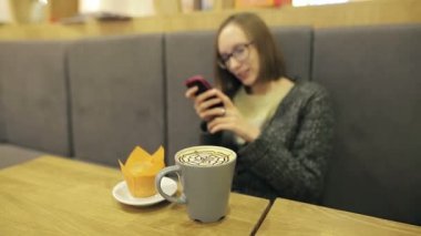 Yalnız Kız bardaklarda kişi sevgilisi için bekliyor. O smartphone cep telefonu üzerinde turuncu kek gülümseyen, mesaj ile Hindistan cevizi kahve içme kafede app kullanma.