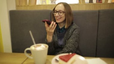 Kadın kanepede otururken speakerphone ile mutlu söz. Gülümseyen ve kameraya bakarak mutlu smartphone Kız bardaklarda hatmi ve kafede, rahatlatıcı ile sıcak çikolata içme .