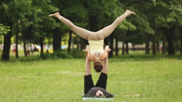 Όμορφο ζευγάρι εξάσκηση acro yoga. Πρακτική εκπαιδευτές νέων γιόγκα σε ένα πάρκο της πόλης για την πράσινη χλόη. Δύο επιτυχημένους νέους εκτελούν ακροβατικά ασκήσεις γιόγκα. Άνδρας και μια γυναίκα να μάθετε γιόγκα στο ηλιοβασίλεμα. — Αρχείο Βίντεο