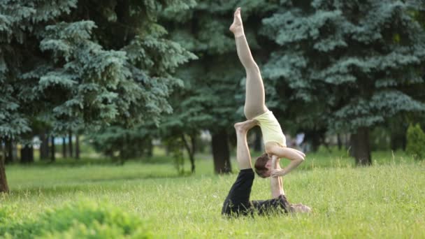 Bella coppia praticare acro yoga. Giovani istruttori di yoga praticano in un parco cittadino su erba verde. Due giovani di successo eseguono esercizi di acro yoga. Uomo e donna imparano lo yoga al tramonto . — Video Stock