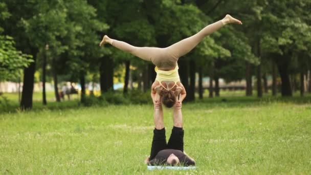 Hermosa pareja practicando acro yoga. Jóvenes instructores de yoga practican en un parque de la ciudad sobre hierba verde. Dos jóvenes exitosos realizan ejercicios de acro yoga. Hombre y una mujer aprenden yoga al atardecer . — Vídeo de stock