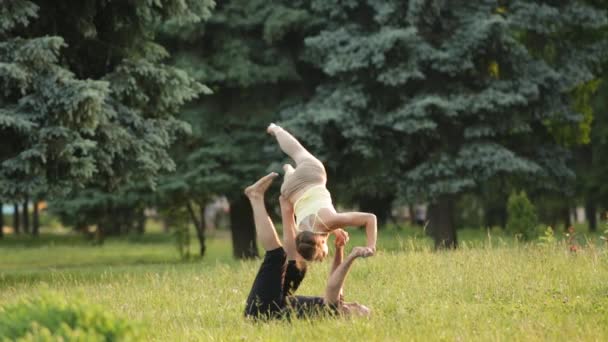 Hermosa pareja practicando acro yoga. Jóvenes instructores de yoga practican en un parque de la ciudad sobre hierba verde. Dos jóvenes exitosos realizan ejercicios de acro yoga. Hombre y una mujer aprenden yoga al atardecer . — Vídeo de stock