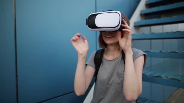 Будущее сейчас. Молодая женщина на лестнице играет в игру в VR-очках. Молодая белая женщина прикасается к чему-то с помощью современных очков виртуальной реальности на синем фоне . — стоковое видео