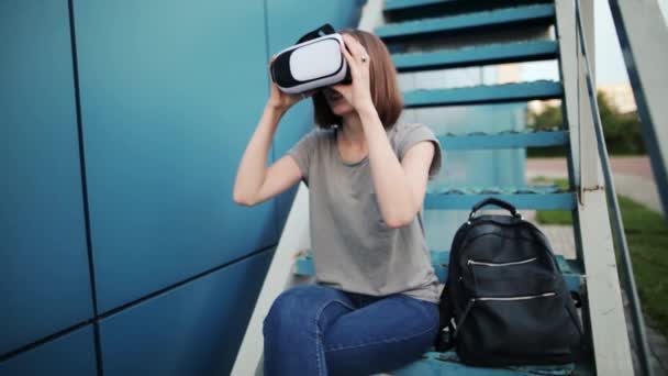 El futuro es ahora. Hermosa joven hembra en una escalera jugando o viendo películas en gafas vr. Joven mujer caucásica tocar algo usando gafas de realidad virtual moderna sobre un fondo azul . — Vídeos de Stock