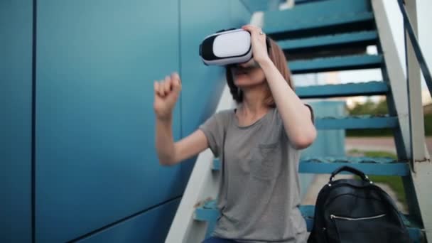 O futuro é agora. Bela jovem fêmea em uma escada jogando jogo em óculos VR. Jovem caucasiana tocar algo usando óculos de realidade virtual moderna em um fundo azul . — Vídeo de Stock