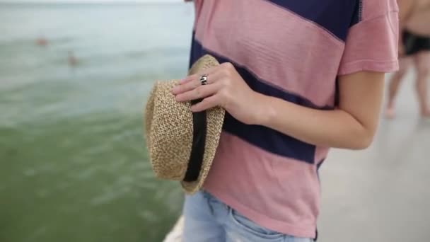 Linda chica o mujer a rayas camiseta y pantalones cortos de mezclilla de pie en el muelle. Mujer sexy está disfrutando del viento en la orilla. Señora está sosteniendo sombrero de sol durante las vacaciones de verano por el mar negro . — Vídeos de Stock