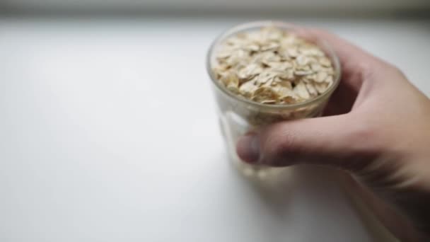 Colazione sana, porridge di farina d'avena. La mano di un uomo caucasico mette il vetro trasparente con grandi fiocchi di farina d'avena su una superficie bianca. Colazione biologica per muscoli forti . — Video Stock