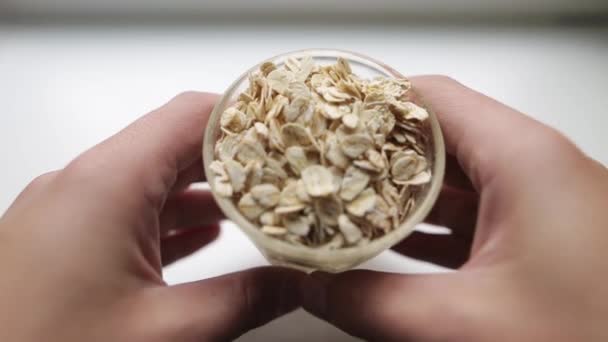 Petit déjeuner sain, porridge aux flocons d'avoine. Deux mains d'un homme caucasien met du verre transparent avec de gros flocons d'avoine sur une surface blanche. Un homme regarde les flocons secs dans une tasse. Aliments biologiques . — Video