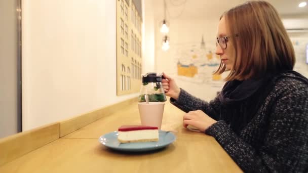 Una donna triste e pensierosa che beve te 'caldo con una cheesecake. La ragazza versa il tè con menta al limone e cannella in una tazza, mescola la bevanda, la sorseggia e guarda fuori dalla finestra, seduta la sera in un caffè. . — Video Stock