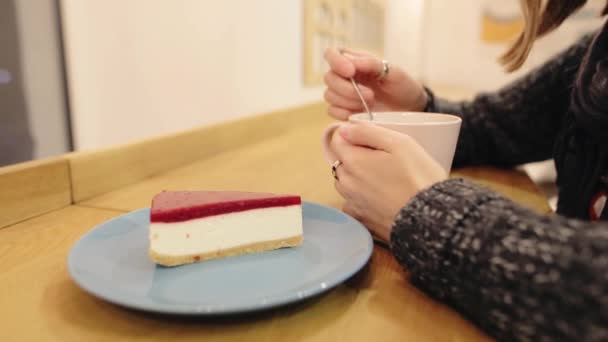 Uma jovem garota atraente mexe chá de hortelã, limão e canela em uma xícara e bebe. uma grande parte de bolo de queijo de morango doce em uma mesa em um café. Hora da noite . — Vídeo de Stock