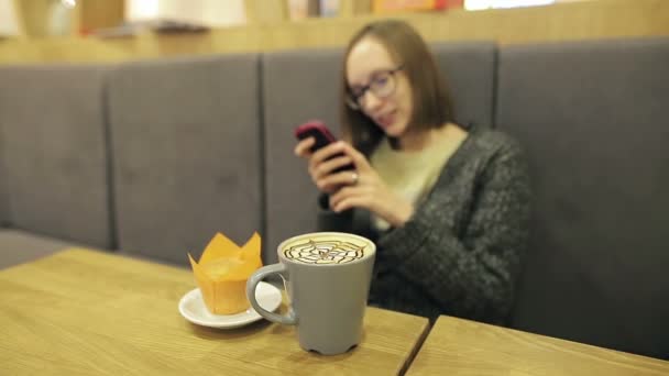 Alone girl in glasses waiting for her boyfriend of other person. She using app on smartphone in cafe drinking coconut coffee with orange cake smiling and texting on mobile phone. — Stock Video