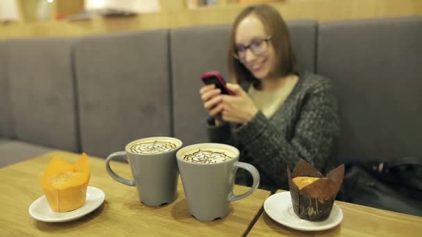 Student girl in glasses surprised using by the text message from her boyfriend. She using app on smartphone in cafe drinking coconut coffee smiling and texting on mobile phone. — Stock Video