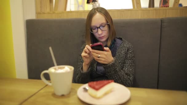 Sad woman using app on smartphone in cafe drinking hot chocolate with marshmallow and texting on mobile phone. while sitting on the sofa. Smartphone girl in glasses. — Stock Video