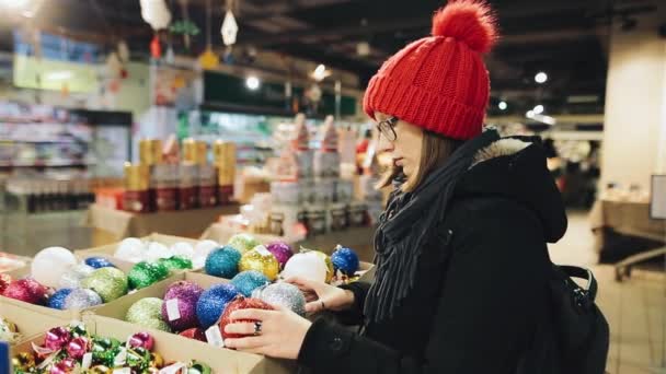 Giovane ragazza caucasica attraente con gli occhiali e un cappello rosso sceglie le palle per l'albero di Capodanno nel negozio del mercato. Una donna sta guardando colorate decorazioni natalizie . — Video Stock