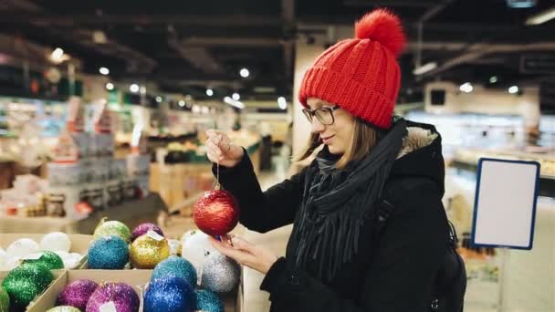 Jolie fille caucasienne en lunettes et un chapeau rouge choisit les boules pour le Noël sur le marché et jouer avec l'un d'eux souriant regardant vers l'appareil photo. Une femme fait pression sur des décorations colorées . — Video
