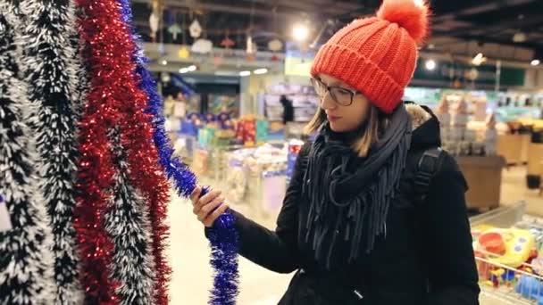 Привлекательная молодая женщина в очках и красной шляпе выбирает рождественский декор для елки. Покупатель нашел дешёвые гирлянды и положил их в корзину. . — стоковое видео