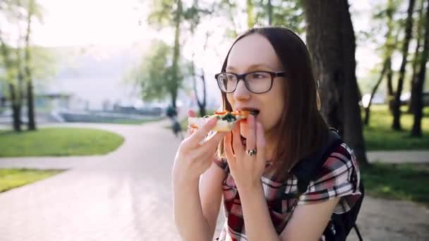 Menina estudante feliz leva uma fatia de pizza, sorri e come sentado em um parque na rua . — Vídeo de Stock