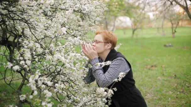 Happy red-haired senior woman looks at and smells cherry tree blossom. Slow motion. — Stock Video