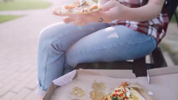 Hipster girl takes a piece of fresh pizza and bites it. The suns rays make their way through her glasses. — Stock Video