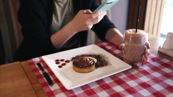 Bir kız şık bir kafede oturmuş akıllı telefonuyla sıcak çikolata içkisinin fotoğrafını çekiyor.. — Stok video